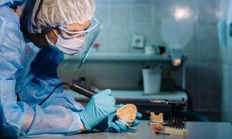 A masked and gloved dental technician works on a prosthetic tooth in his lab photo