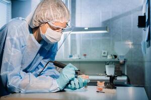 A masked and gloved dental technician works on a prosthetic tooth in his lab photo