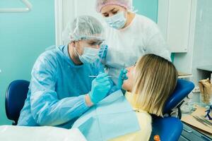 a dentist in a protective mask sits next to him and treats a patient in the dental office with an assistant photo