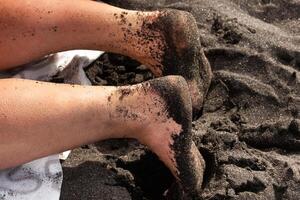 de cerca de un mujer piernas y pies en el arena, acostado en el volcánico negro arena en el playa.tenerife foto