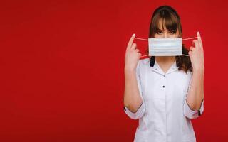 A female doctor stands on a red background and holds a medical mask in her hands photo