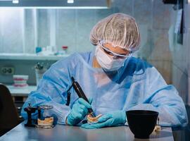 A masked and gloved dental technician works on a prosthetic tooth in his lab photo