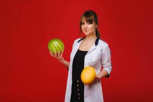 a female doctor nurse in a white coat with fruit in her hands poses on a red background, melon, watermelon, photo