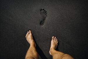 cerca arriba de masculino huellas y pies caminando en el volcánico negro arena en el playa foto