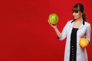 a female doctor nurse in a white coat with fruit in her hands poses on a red background, melon, watermelon, photo