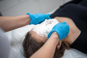 Doctor cosmetologist or dermatologist making face mask in cosmetology salon. Professional Beautician applying face mask on caucasian woman face lying on bed in bathrobe. photo