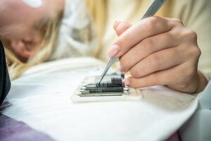 The master takes a bunch of curved artificial eyelashes with tweezers from the tablet and dips into the glue. The process of eyelash extension in a beauty salon. photo