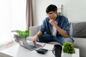 el hombre en casual ropa trabajando con un computadora portátil, computadora, inteligente teléfono, calculadora sentado en el sofá en el vivo habitación a hogar, trabajando desde hogar concepto. foto