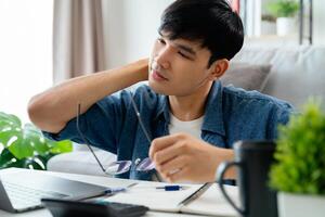 A tired man wearing glasses is sitting at a desk with a laptop and a notebook photo