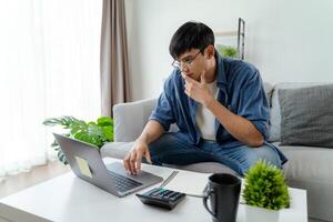 el hombre en casual ropa trabajando con un computadora portátil, computadora, inteligente teléfono, calculadora sentado en el sofá en el vivo habitación a hogar, trabajando desde hogar concepto. foto
