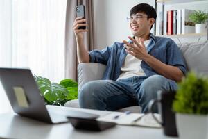 alegre contento joven asiático hombre utilizando inteligente teléfono para en línea vídeo conferencia llamada hablando felizmente. foto