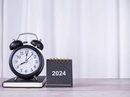 creativo escritorio con 2024 calendario, negro alarma reloj en de tapa dura libro con blanco cortina para antecedentes. Copiar espacio para texto, oficina escritorio concepto. foto