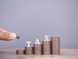 Close up hand putting coin into stack of coins and Wooden words with the TAX. The concept about saving money and manage time to paying tax photo