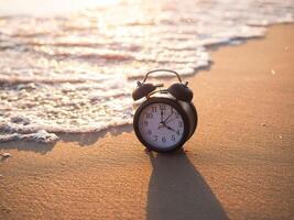 negro alarma reloj en el playa en el puesta de sol tiempo. el concepto acerca de hora a verano, viajar, vacaciones y relajación foto