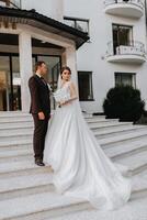 Groom and bride walking outdoors near a posh hotel. A long train of dress on the steps photo