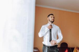 a young man in a white shirt stands by the window in the room and puts on a tie. The groom gets dressed in the morning and prepares for the wedding. photo