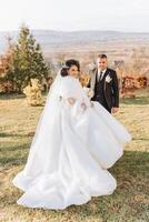 un Boda Pareja es caminando en naturaleza en un otoño día. contento joven novia y elegante novio participación manos. un elegante Pareja de recién casados en su Boda día. foto