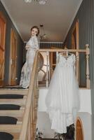 A delicate wedding dress hangs on a beautiful railing in the hallway of the house. The bride climbs the steps in a delicate transparent petticoat. Nice light. Wedding photo. A high quality photo
