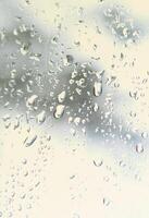 A photo of rain drops on the window glass with a blurred view of the blossoming green trees. Abstract image showing cloudy and rainy weather conditions