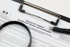 IRS Form W-9 Request for taxpayer identification number and certification blank on A4 tablet lies on office table with pen and magnifying glass photo