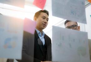 Two men are looking at a wall covered in paper with graphs and charts. They are discussing the information on the wall photo
