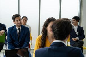 A group of people are sitting in a room, some of them wearing suits. A man in a suit is talking to a woman in a yellow dress. Scene is professional and formal photo