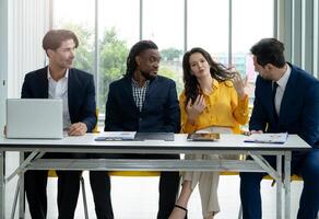 A group of people are sitting around a table in a business setting. One woman is talking to the group while the others listen. The atmosphere is professional and serious photo