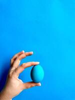 Hand balancing blue egg on a fingertip against solid blue background, minimalist concept for balance, Easter, and simplicity with space for text. photo