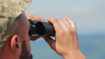 A man in a camouflage cap looks through binoculars. Close-up side view. The soldier examines enemy positions through binoculars. video