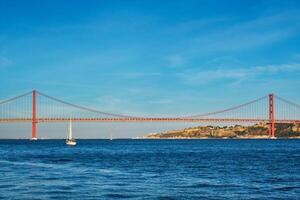 View of 25 de Abril Bridge over Tagus river, Christ the King monument and a yacht boat at sunset. Lisbon, Portugal photo