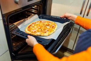A woman taking out baking tray with delicious pizza from oven on kitchen photo