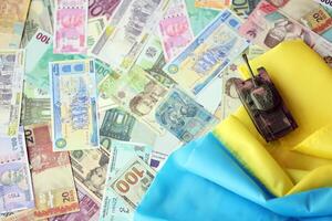 Toy tank on Ukrainian flag on many banknotes of different currency. Background of war funding photo