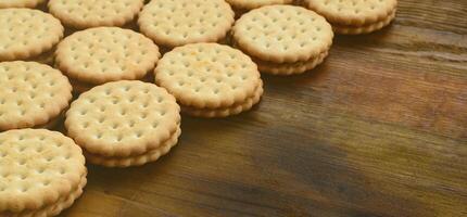 A round sandwich cookie with coconut filling lies in large quantities on a brown wooden surface. Photo of edible treats on a wooden background with copy space