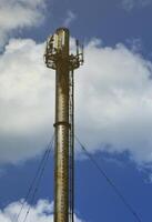 High metal chimney of industrial plant with ladder in the form of metal braces against the background of a cloudy blue sky photo