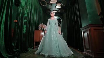 Elegant woman in a sparkling gown standing on stage with dramatic lighting and green curtains in the background. video