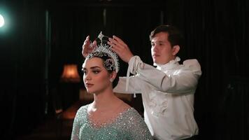 Elegant woman in a sparkling gown with a crown being adjusted by a man on a dark stage with dramatic lighting. video