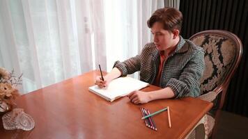 Man in a tweed jacket writing in a notebook at a wooden table, with pencils and a flower vase, near a window with sheer curtains. video