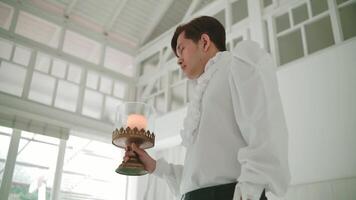 Serene man in white holding a lit candle in a tranquil room with large windows. video