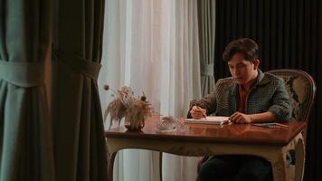 Focused individual writing in a notebook at a wooden table, with soft lighting and curtains video