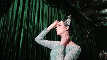 Elegant woman in sparkling dress posing with hand on forehead against a green curtain backdrop, vintage style. video