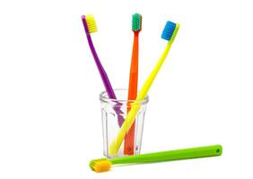 Three toothbrushes - purple, orange and yellow with multi-coloured bristles in a transparent cup on a white background. The green brush lies next to the cup. Isolated. Vertical. High quality photo. photo