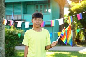 Asian boy holds a rainbow flag in front of a house decorated with rainbow flags during Pride Month to show LGBT pride and identity. Soft and selective focus. photo