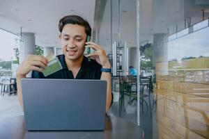 Internet compras. joven asiático hombre pago para en línea compra, utilizando crédito tarjeta, célula teléfono y computadora portátil, sentado a cafetería. foto