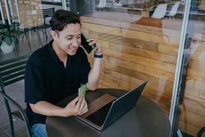 Internet compras. joven asiático hombre pago para en línea compra, utilizando crédito tarjeta, célula teléfono y computadora portátil, sentado a cafetería. foto