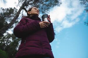 fondo vista. viajero mujer participación un termo taza con caliente beber, té o café, mirando lejos, excursionismo en el bosque foto