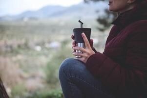 Female traveler holding thermos mug with hot drink, warming hands while relaxing after hiking in the forest or mountains photo