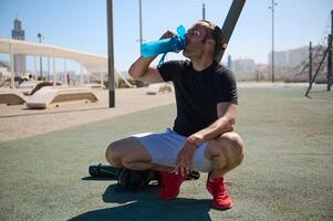 activo atlético joven hombre Bebiendo agua desde Deportes botella mientras descansando en el campo deportivo después rutina de ejercicio al aire libre foto