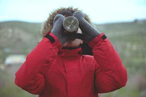 Teenager boy in red parka with fluffy further hood, drinking from steel travel thermos mug against mountains background photo