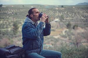 masculino viajero aventurero con mochila, sentado en un Iniciar sesión en el bosque, Bebiendo café desde un termo jarra foto