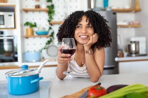 Smiling young housewife with red wine glass looking at the camera as she stands at the stove in the kitchen preparing dinner photo
